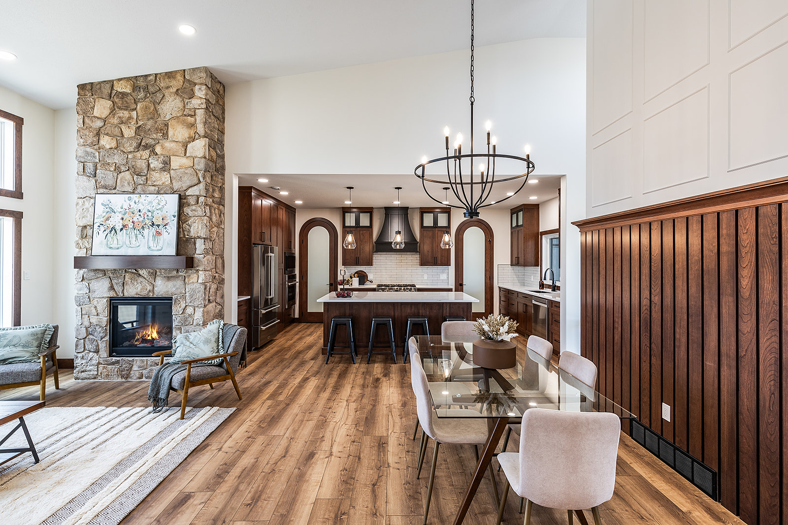 Spacious kitchen and dining area with wooden floors. Stone fireplace on the left, modern kitchen island with stools, glass table, and minimalist decor. Warm and inviting.