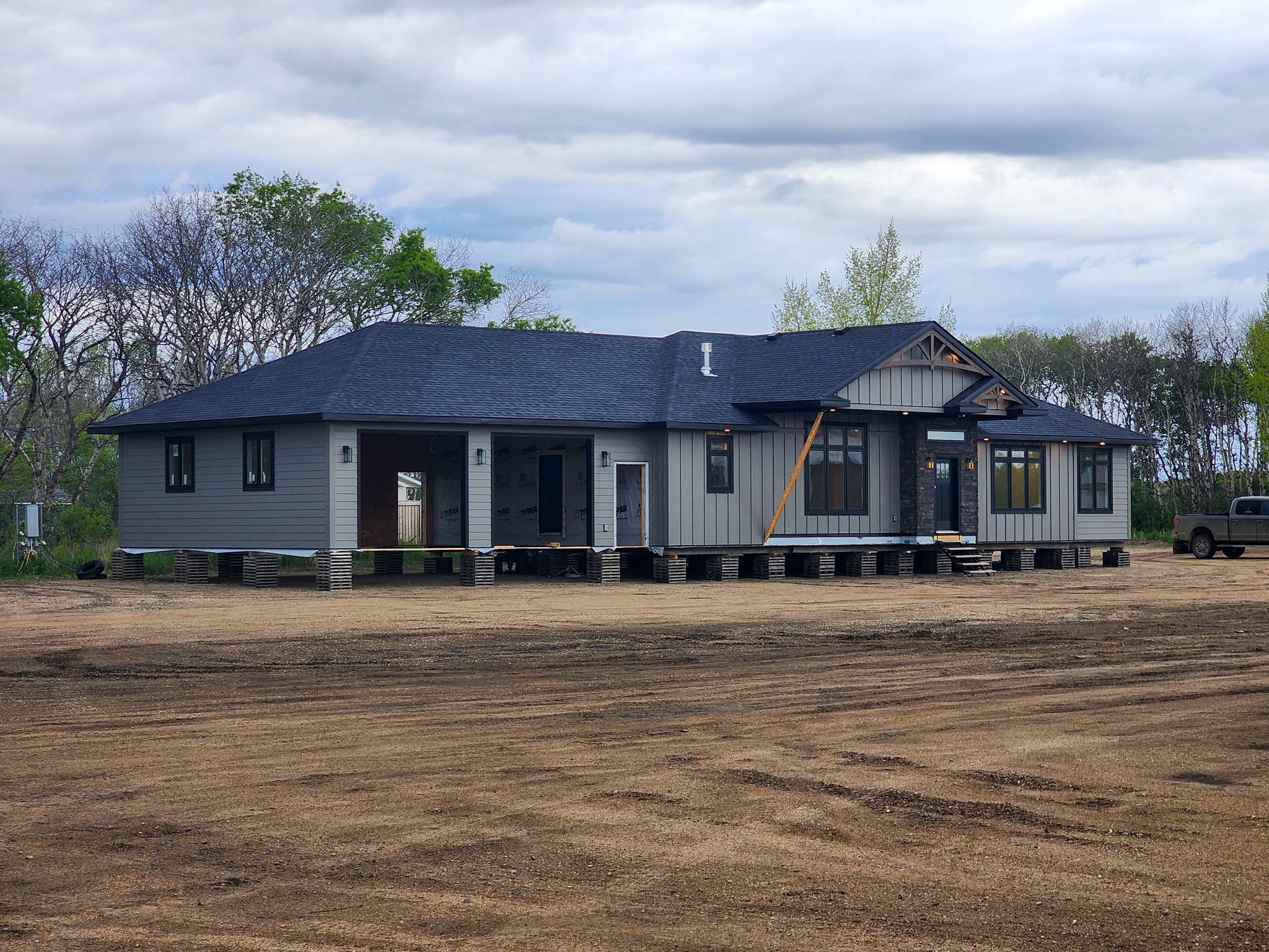 A single-story house under construction stands elevated on a dirt plot, surrounded by sparse trees under a cloudy sky, creating a calm and serene atmosphere.