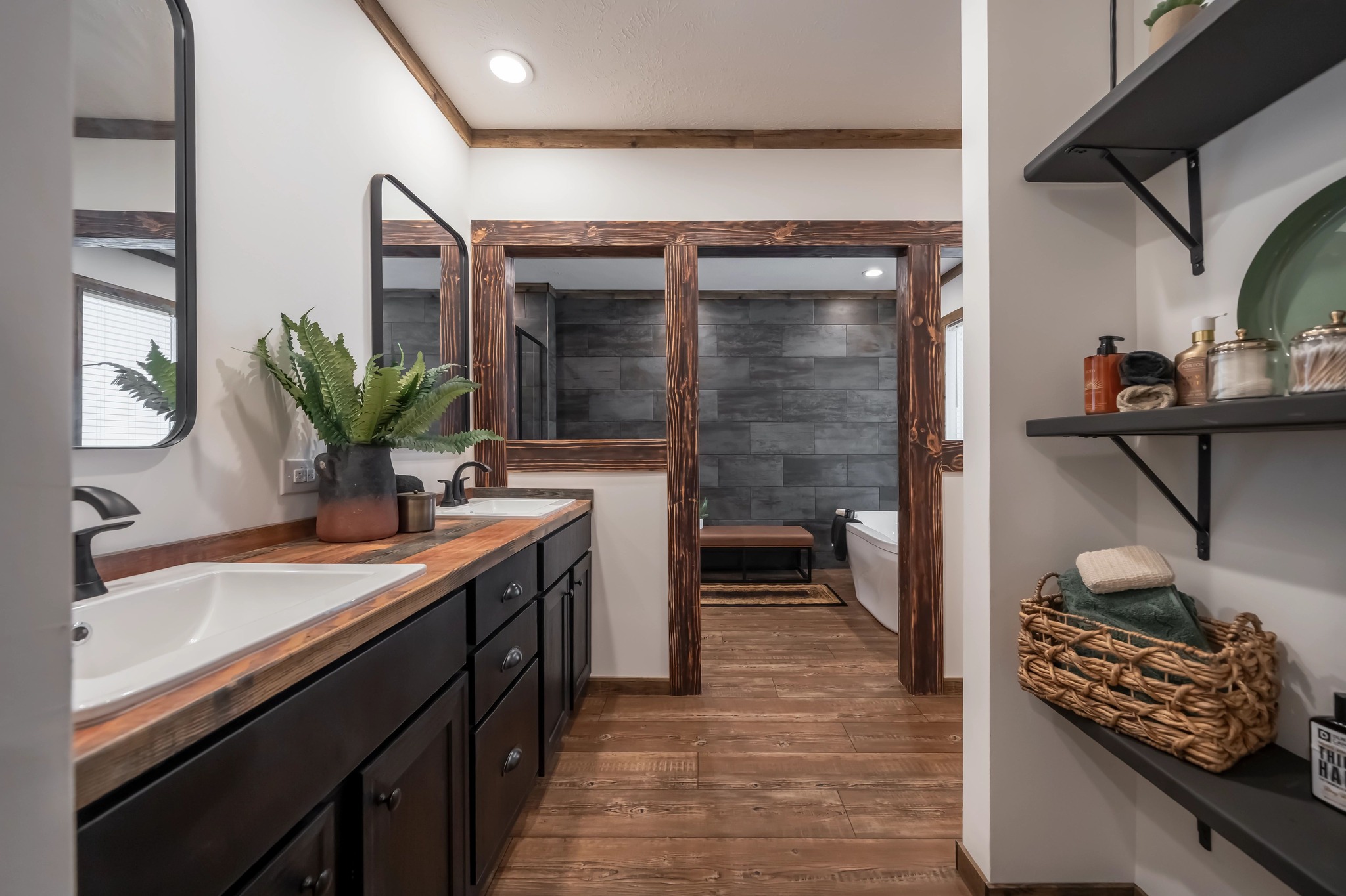 A rustic bathroom features a dual-sink vanity with dark wood cabinets and a wood countertop. Plants add greenery. Open shelves display toiletries. The gray stone wall and wood accents create a warm, serene ambiance.