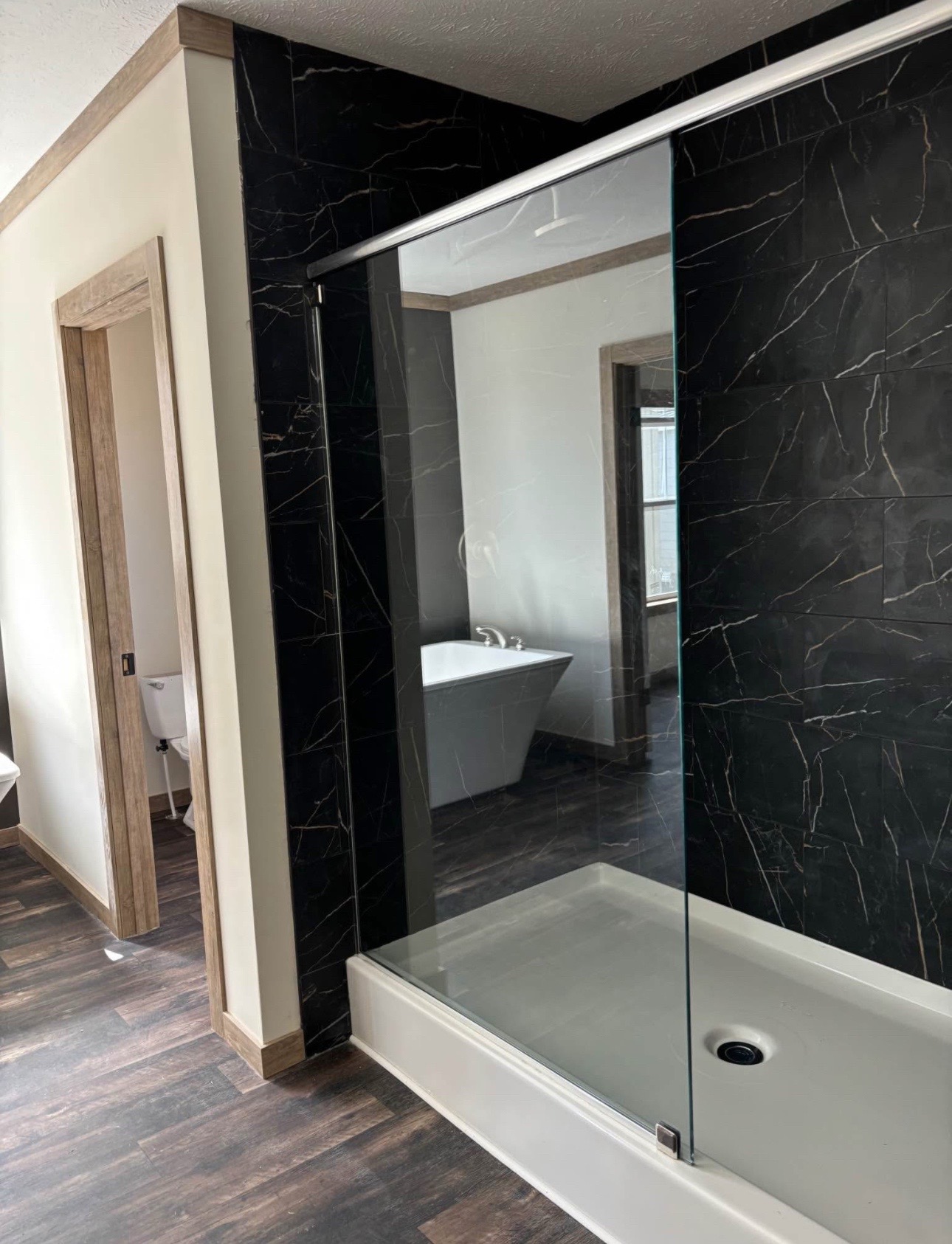 Modern bathroom with dark marble walls, a glass shower enclosure, and a white bathtub. Wooden floor and trim add warmth and contrast.