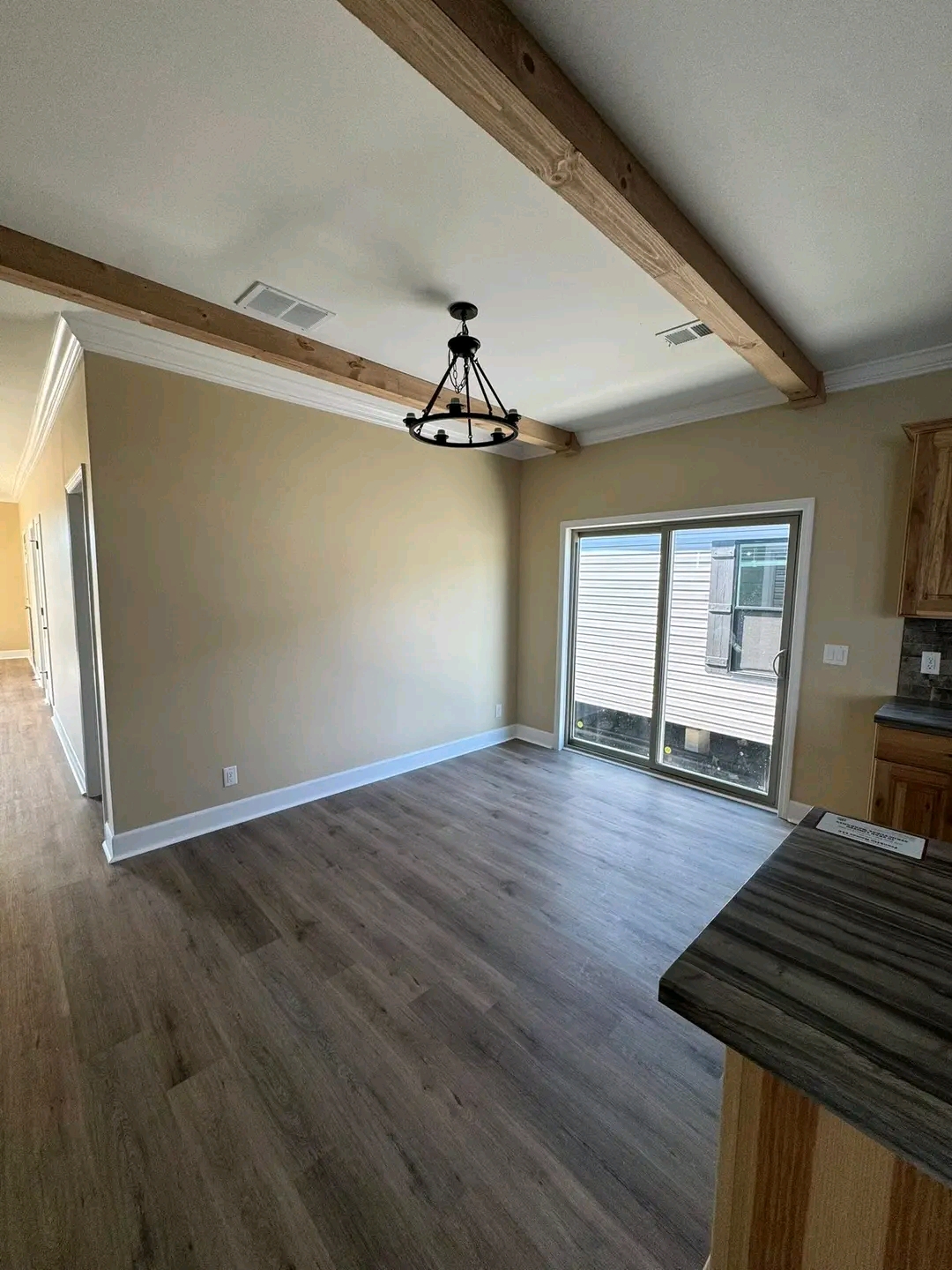 Modern, empty room with light wooden flooring, beige walls, and wooden ceiling beams. A black chandelier hangs from the ceiling. Sliding glass doors allow natural light.
