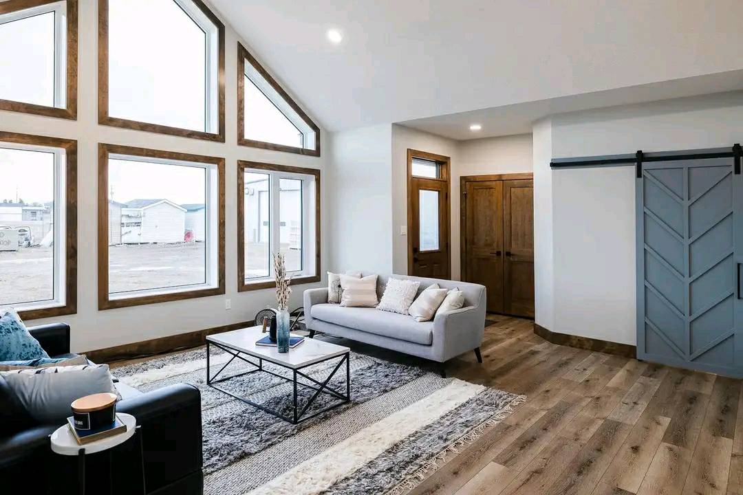 Spacious living room with large, triangular windows and wooden frames, featuring a gray sofa, black armchair, and a modern coffee table on a striped rug.