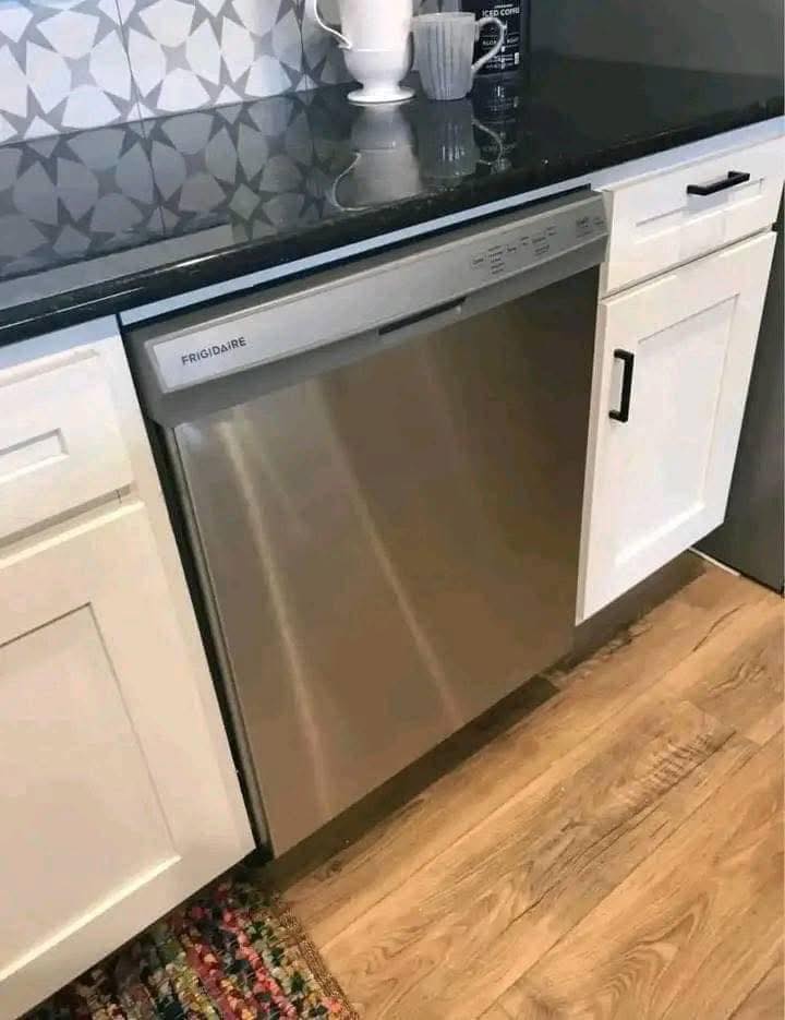 Modern kitchen with a stainless steel dishwasher, black countertop, geometric tiled backsplash, and white cabinets on a wooden floor, creating a sleek, clean look.