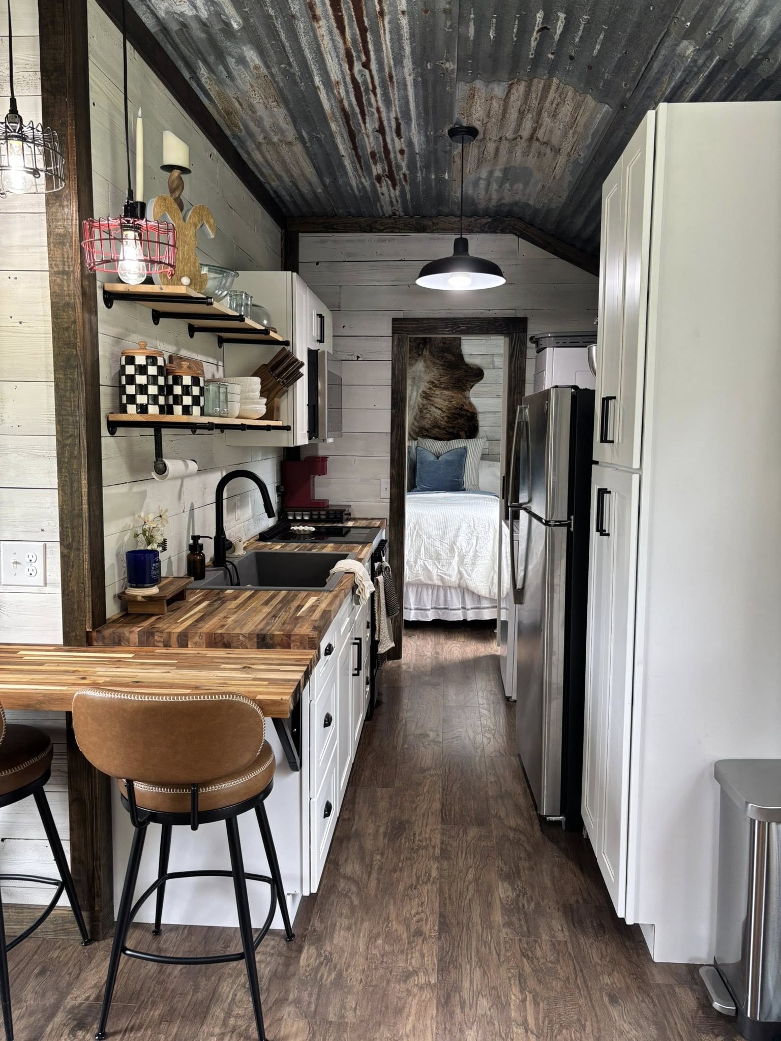 Narrow kitchen with wood countertops, open shelves, and a corrugated metal ceiling. A cozy bedroom is visible at the end, conveying a rustic, intimate feel.