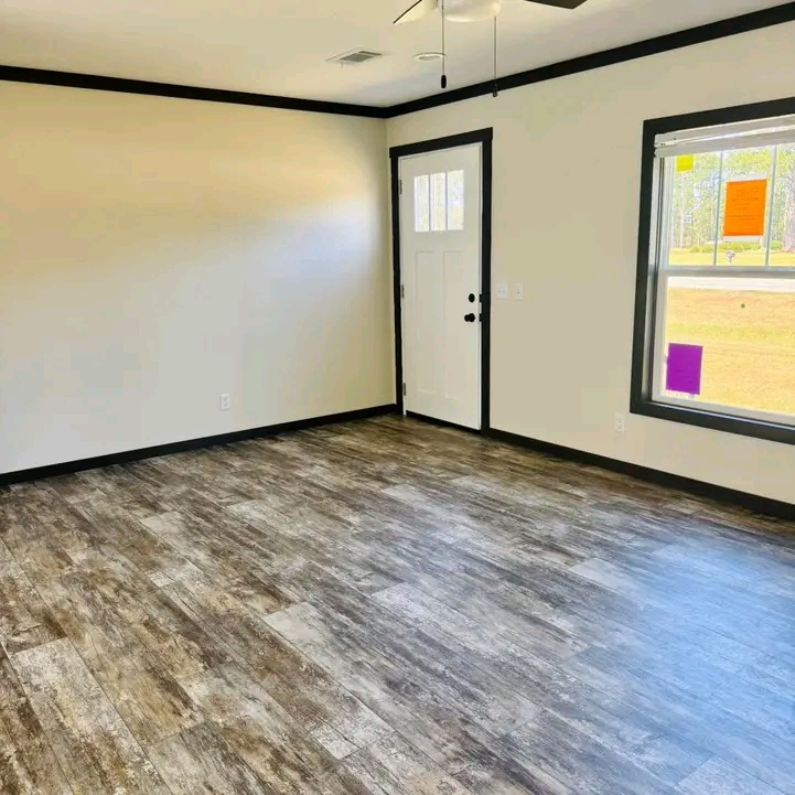 Empty room with light beige walls, dark wood-look flooring, and a white door with black trim. Sunlight streams through a large window, creating a warm, inviting atmosphere.