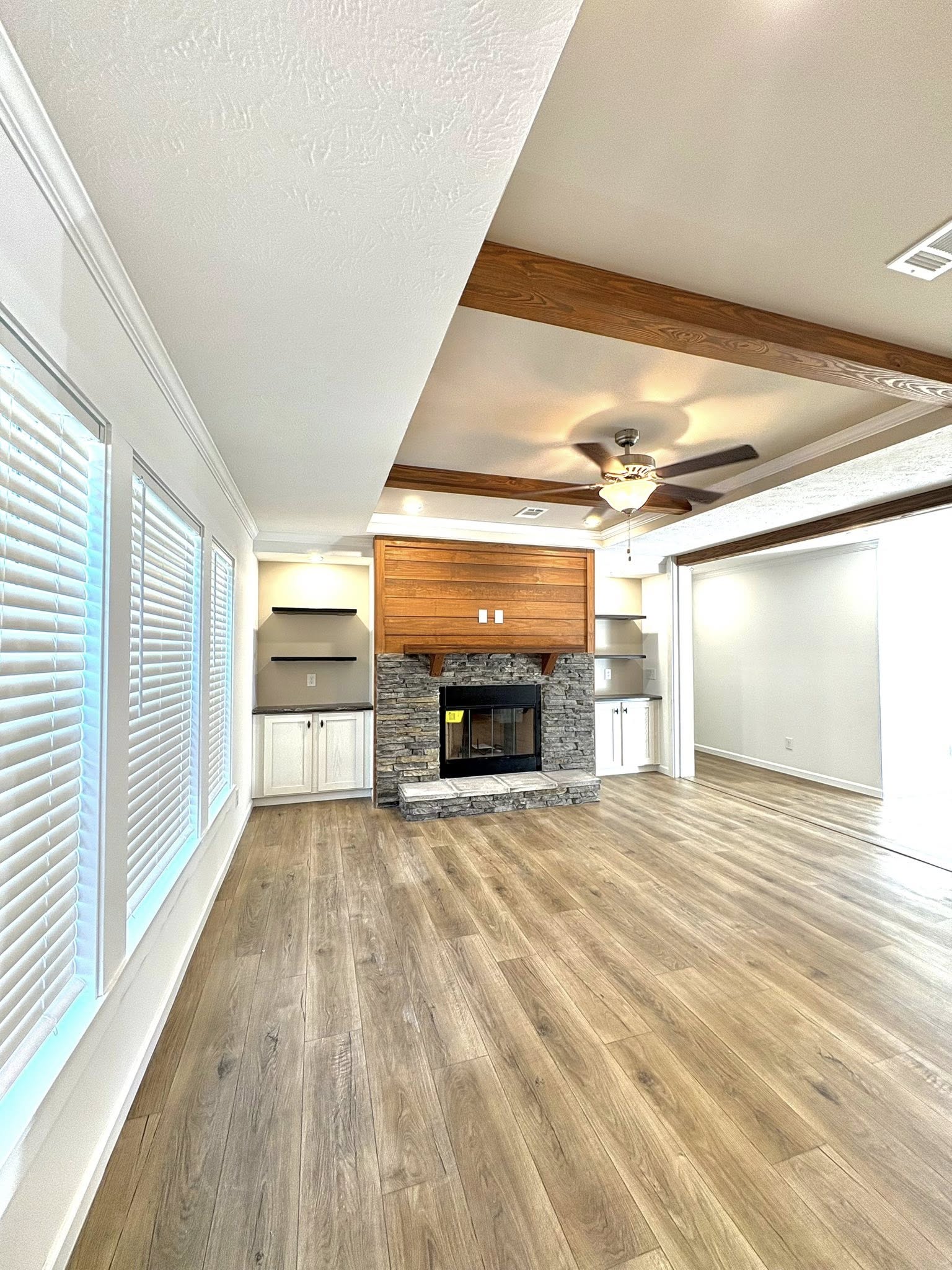 Spacious living room with wood floors, an elegant stone fireplace, and ceiling beams. Soft natural light through large windows creates a cozy atmosphere.