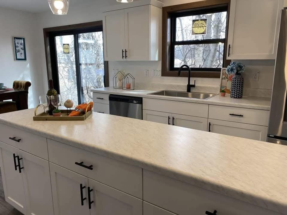 Modern kitchen with white cabinets and black handles, a wide marble countertop, and stainless steel appliances. A tray with wine, vegetables, and a vase with flowers adds warmth. Large window and sliding doors offer natural light, creating an inviting space.