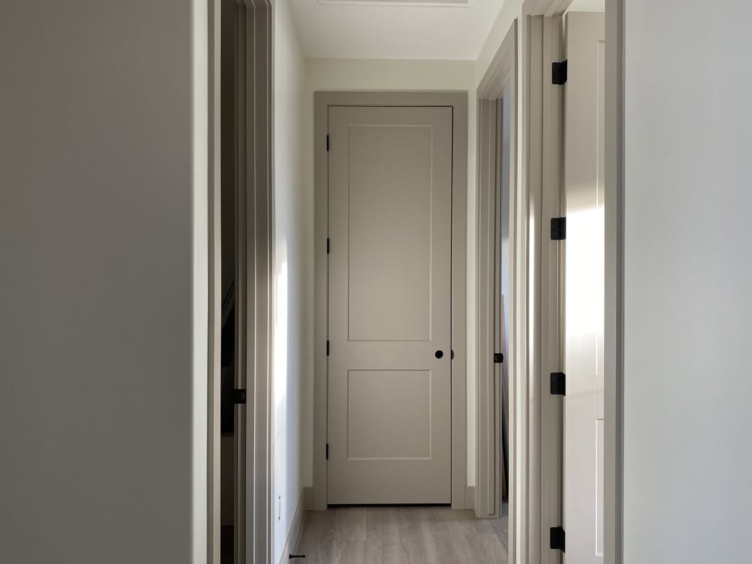 View of a narrow hallway with light wooden flooring, flanked by closed beige doors on both sides. Soft, natural lighting creates a calm ambiance.
