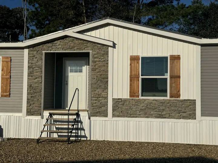 Front of a modular home with beige siding, stone accents, a small porch, steps leading up, and windows with wooden shutters. Trees in the background.