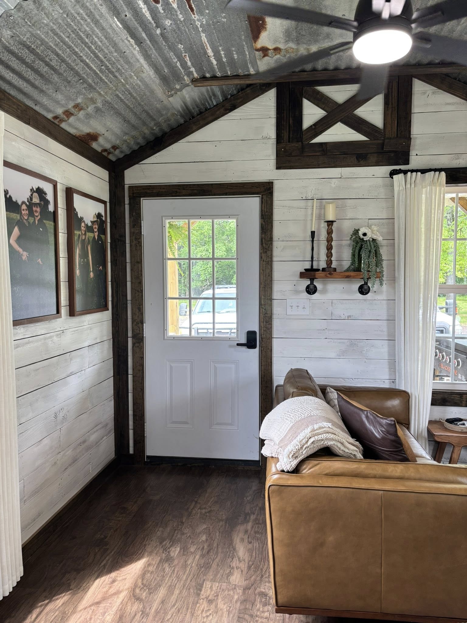 Cozy rustic room with white shiplap walls and a corrugated metal ceiling. Features a brown leather sofa, decorative shelf, photos, and a windowed door. Warm ambiance.