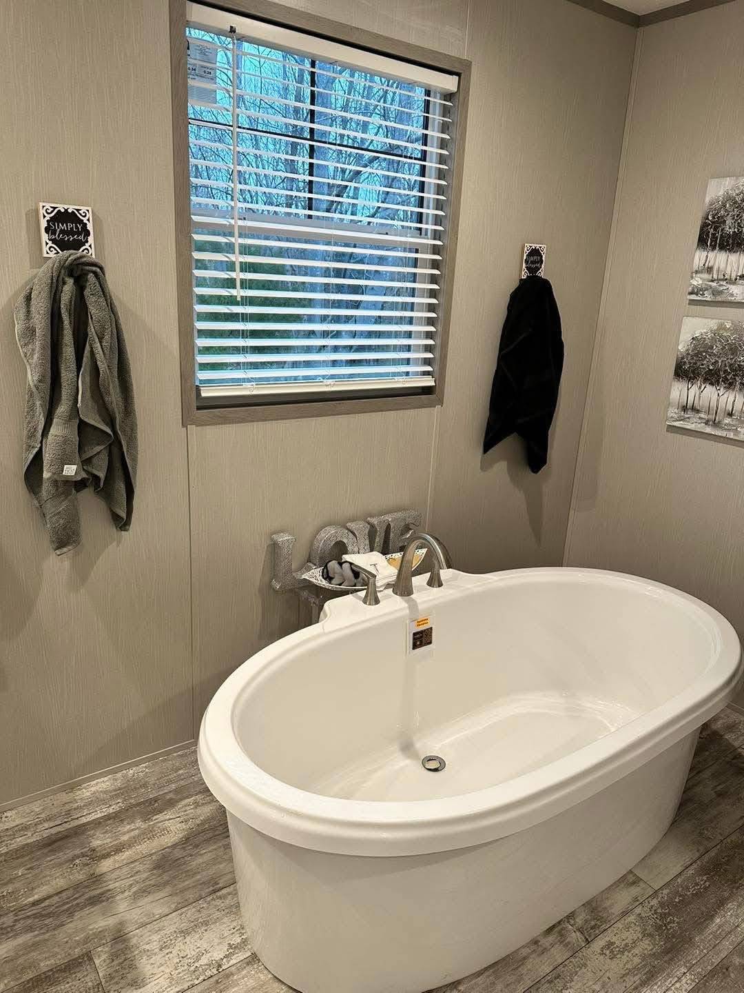 A modern bathroom features an oval bathtub with silver faucets, gray walls, a window with blinds, two towels, and a serene, minimalist decor.