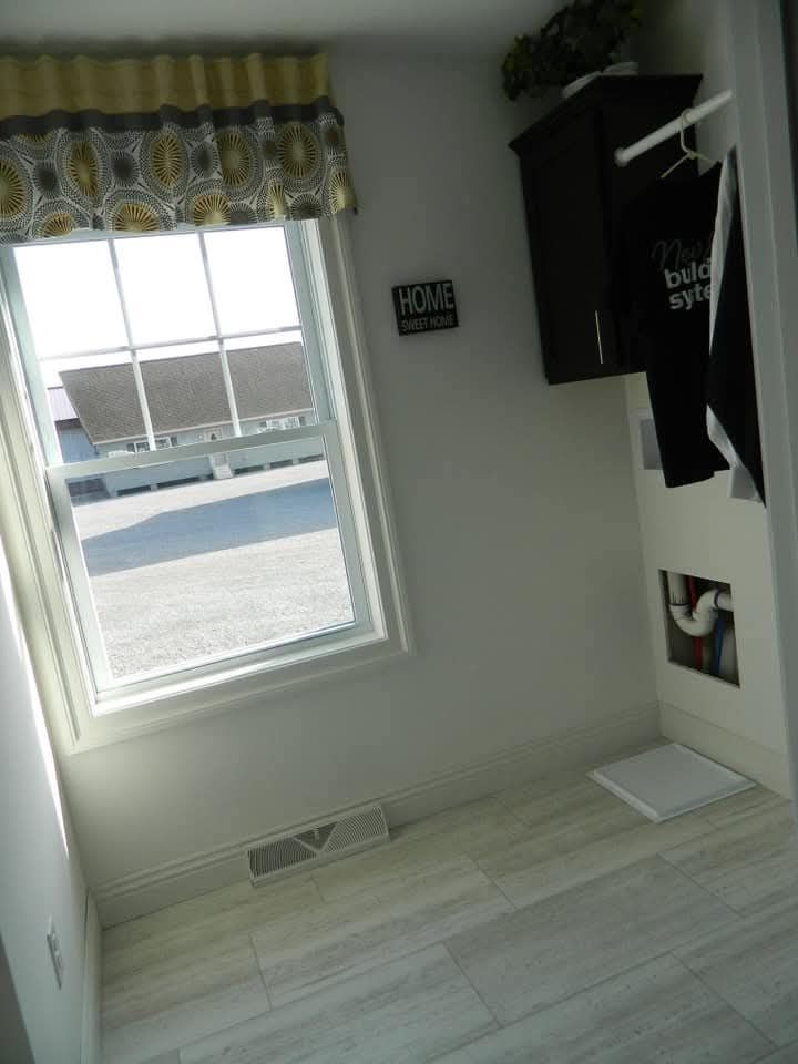 A bright, minimalist laundry room with a large window, patterned curtains, a "Home Sweet Home" sign, and light tile flooring. Sunlight streams in.