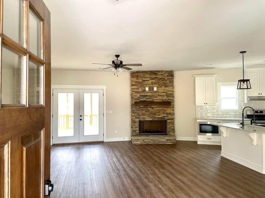 Spacious living area with wood flooring, featuring a stone fireplace, ceiling fan, and modern kitchen with white cabinets. French doors lead outside.