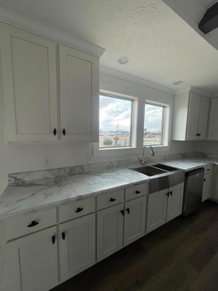 Bright kitchen with white cabinets, marble countertops, and a stainless steel sink. Large windows provide natural light and a view outside. Calm and modern ambiance.