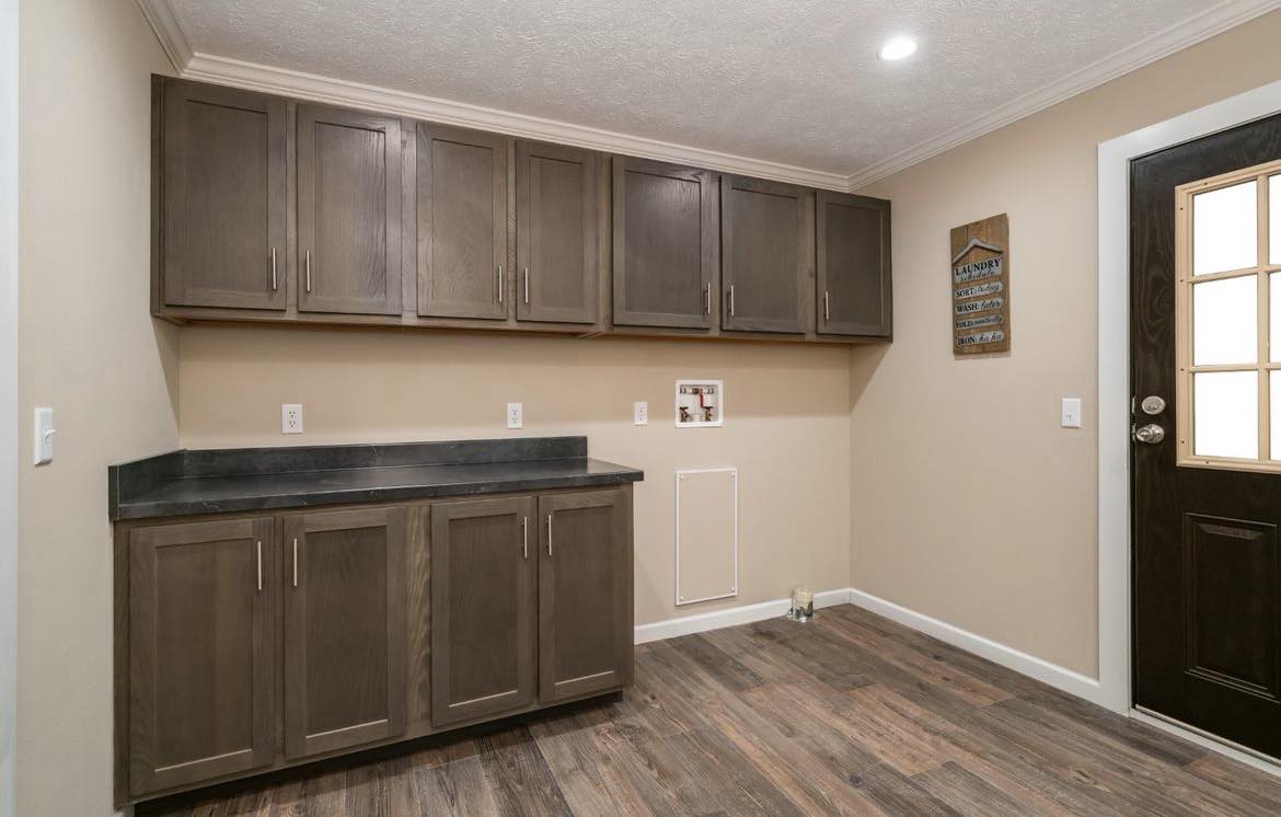 A tidy room with dark wooden cabinets, both upper and lower, against beige walls. A black countertop and a dark wooden door create a warm, modern look.