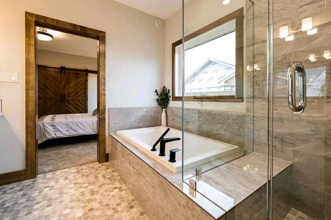 Modern bathroom with a glass shower, beige marble bathtub, and black fixtures. A window overlooks a house. Barn door leads to a bedroom.