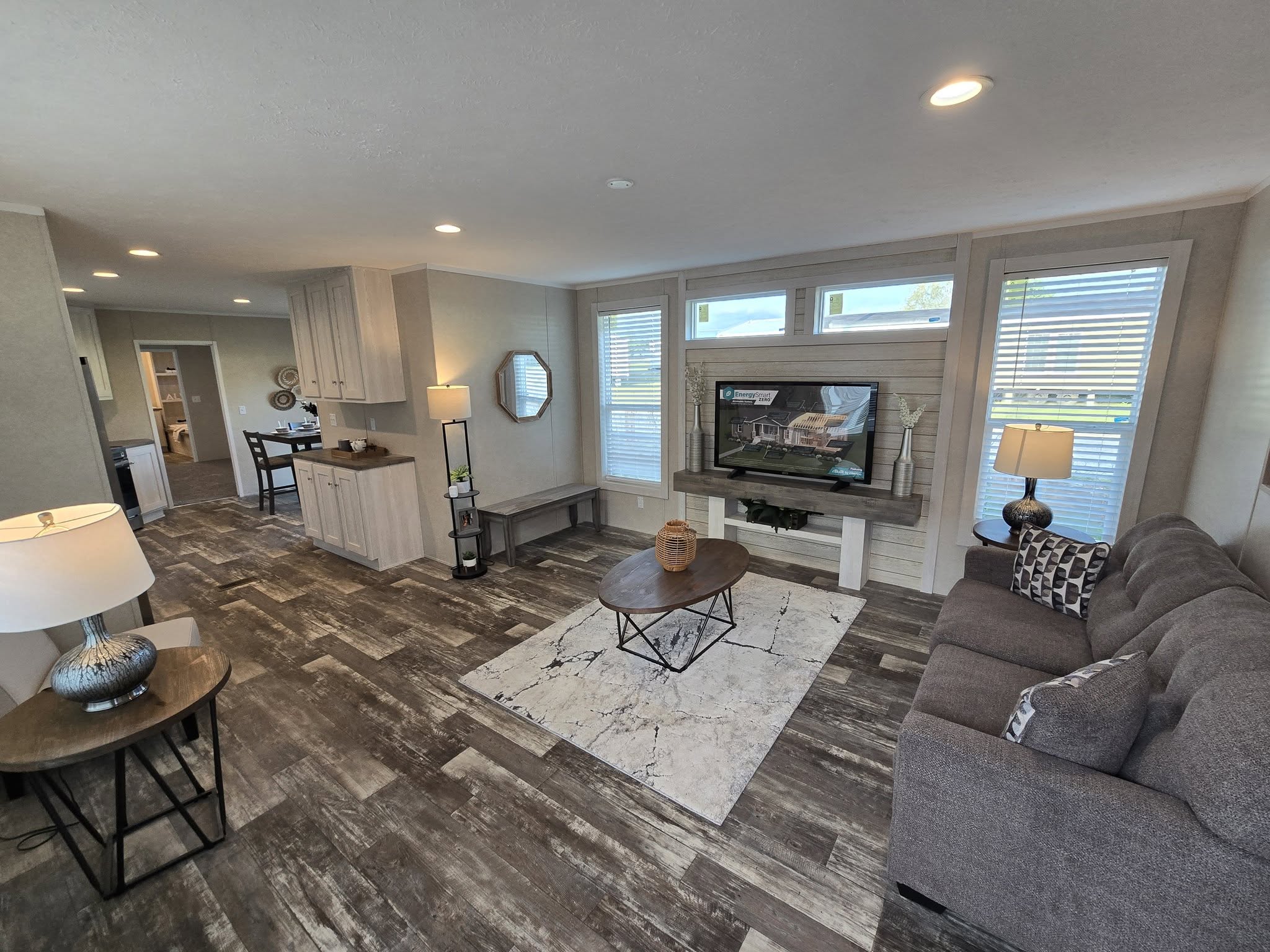 Spacious modern living room with a gray sofa, wooden coffee table on a textured rug, and a large TV. Bright natural light streams through tall windows.