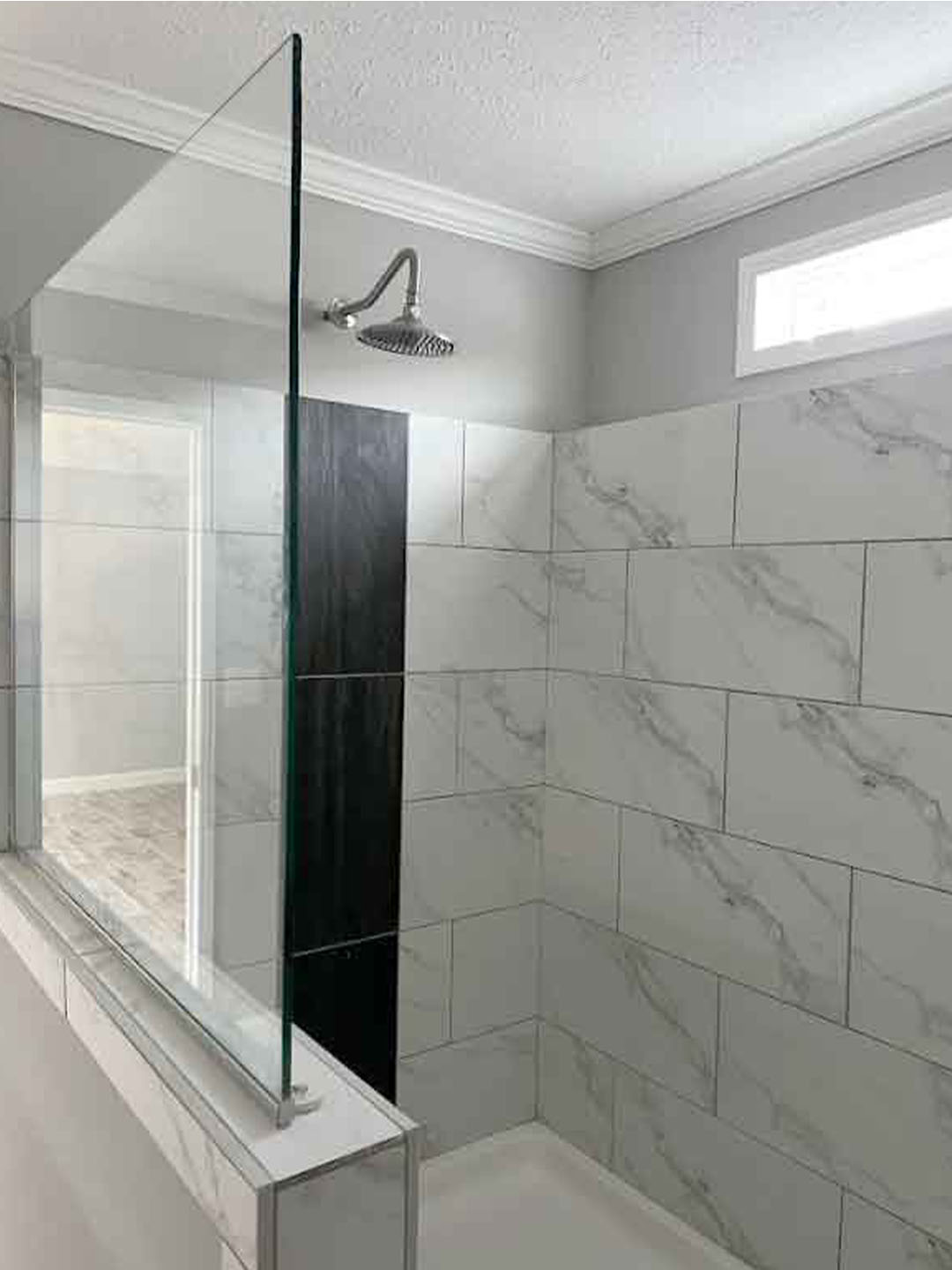 Modern bathroom with gray marble tiles and a rain showerhead. A glass partition separates the shower area, creating a clean, minimalist look. Light fills the space.