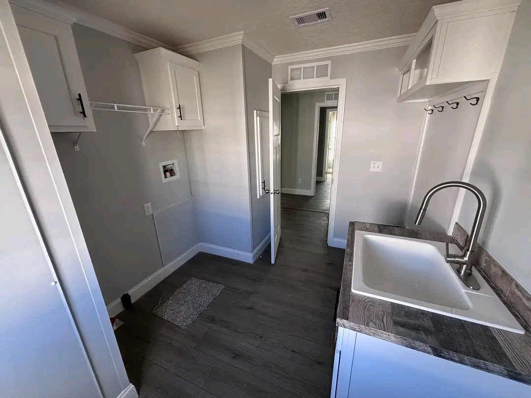 Bright, modern laundry room with light gray walls, wooden floor, and a white sink on a dark countertop. Cabinets and open door create a spacious feel.