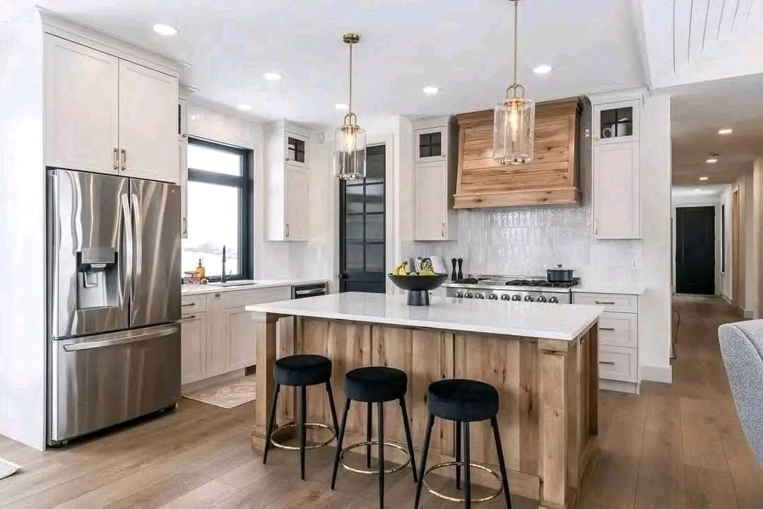 Modern kitchen with a central island featuring wood paneling and three black stools. Stainless steel appliances and pendant lights convey a sleek, cozy vibe.
