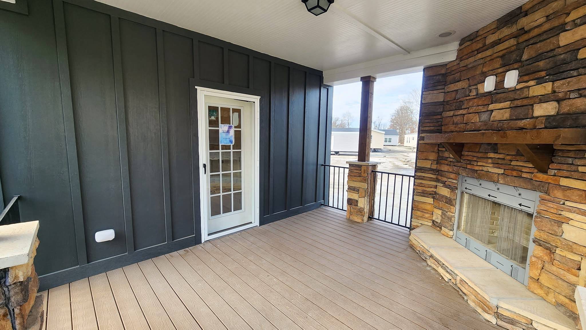 Covered patio with a rustic stone fireplace and dark-paneled wall. Light wood flooring contrasts with a white-framed door leading outside. Bright and inviting.