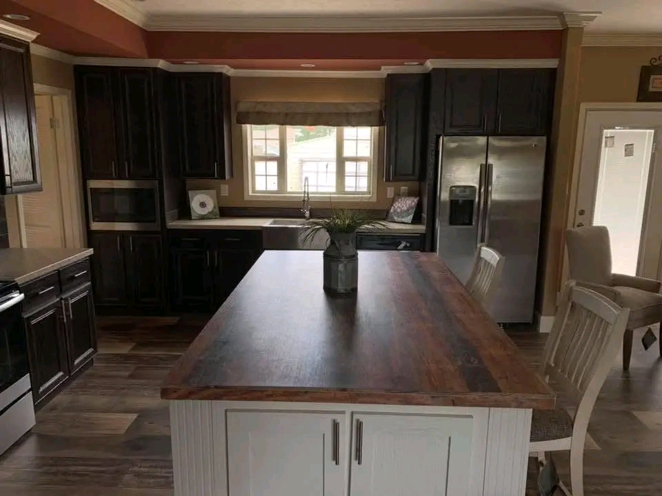 A modern kitchen with dark cabinets, a stainless steel fridge, and a large island with a wooden countertop. Sunlight streams through a window, creating a warm atmosphere.