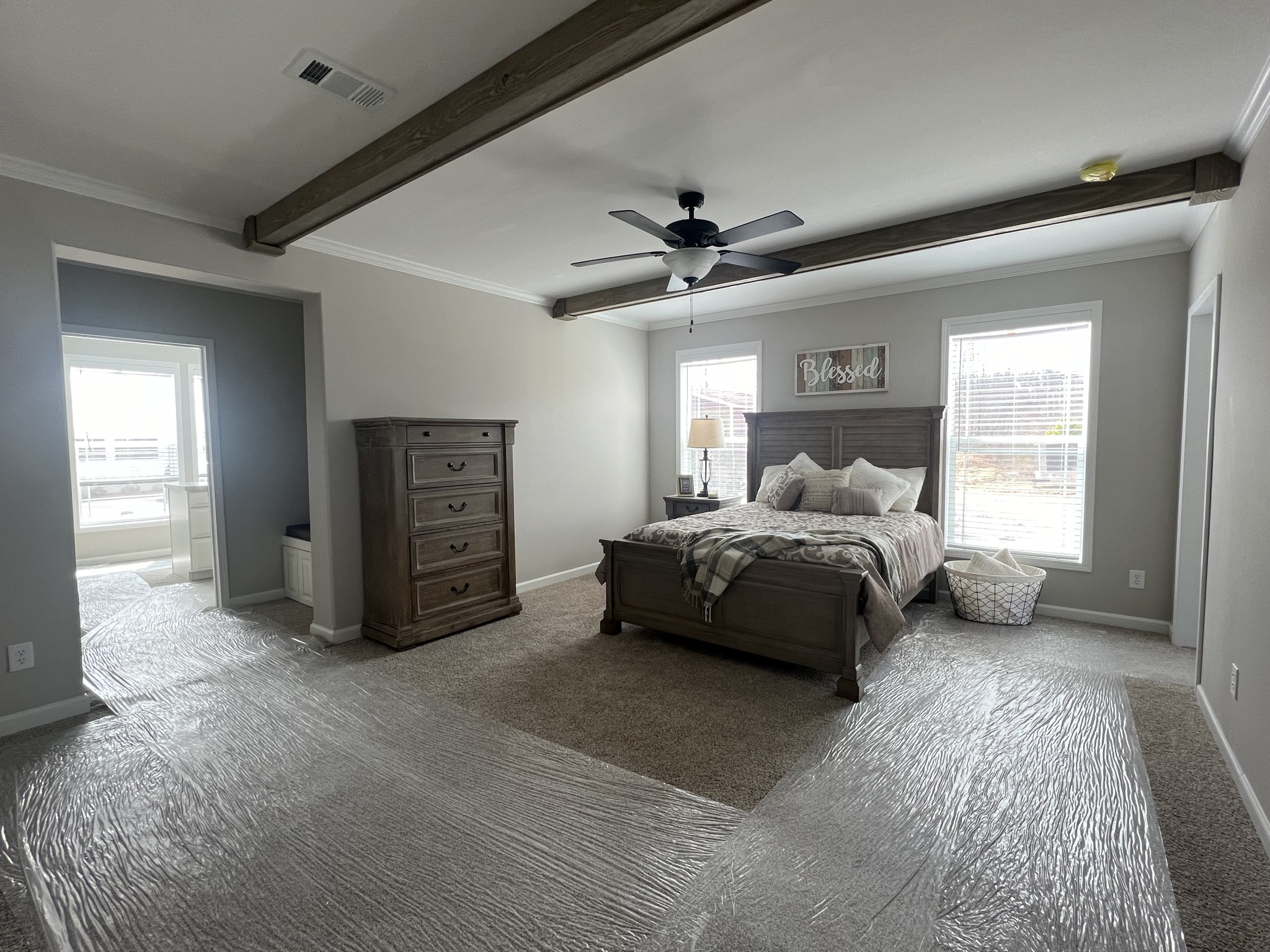 Spacious bedroom with a cozy feel, featuring a wooden bed, plush bedding, large dresser, ceiling fan, and sunlight streaming through two windows.
