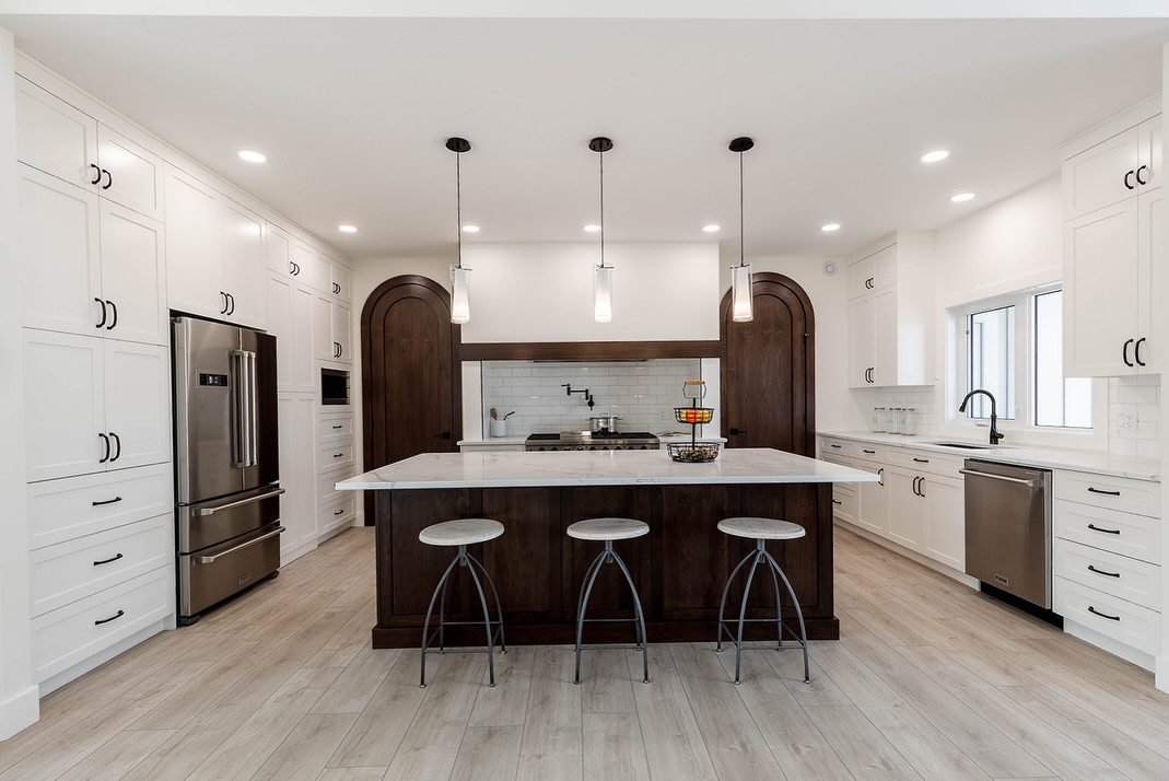 Sleek modern kitchen with a large island, pendant lights, and bar stools. Features white cabinets, stainless steel appliances, and wood accents.