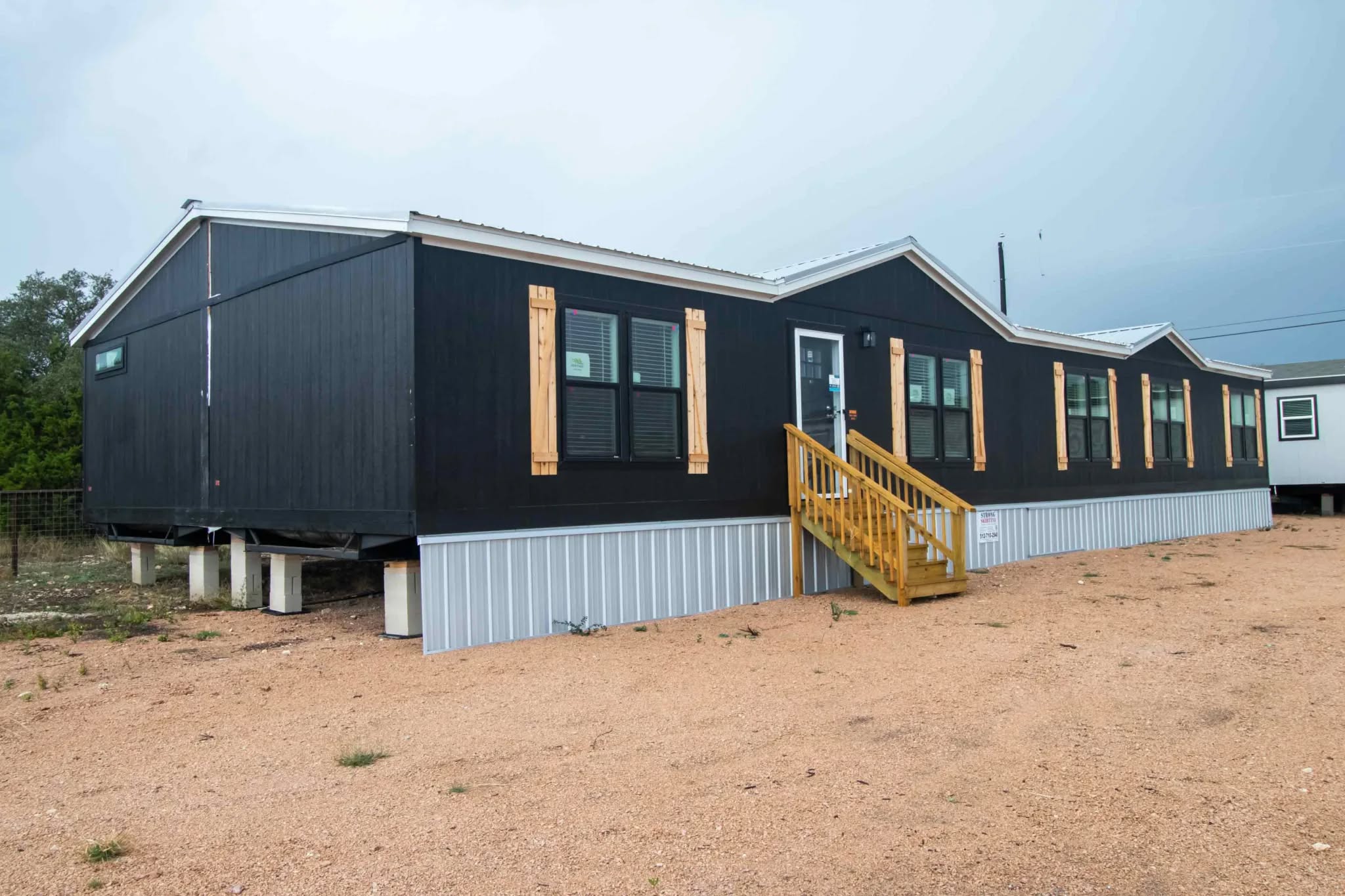 Long, dark-colored modular home on raised foundation with multiple windows, wooden shutters, and a small staircase leading to the entrance. Overcast sky above.