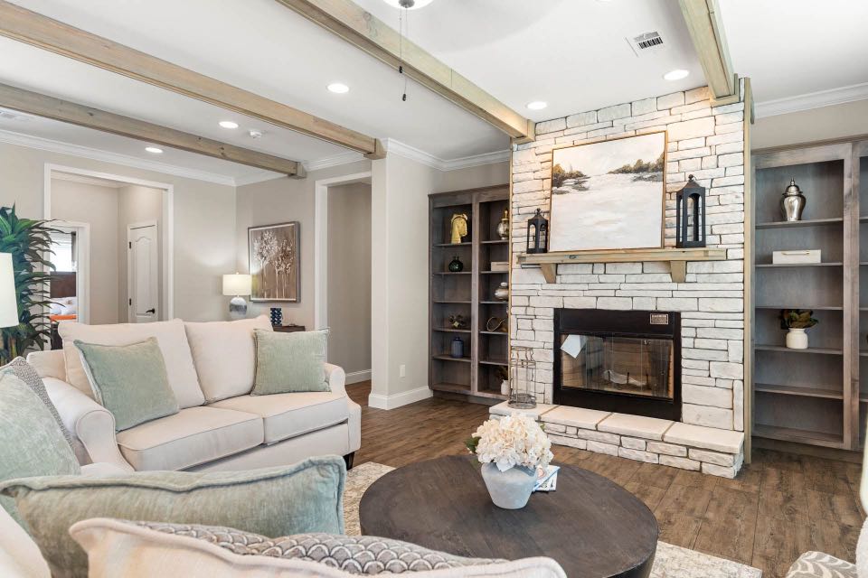 Chic living room with cream sofas, soft green cushions, wooden beams, stone fireplace, art above mantel, shelves with decor, and a warm, cozy ambiance.