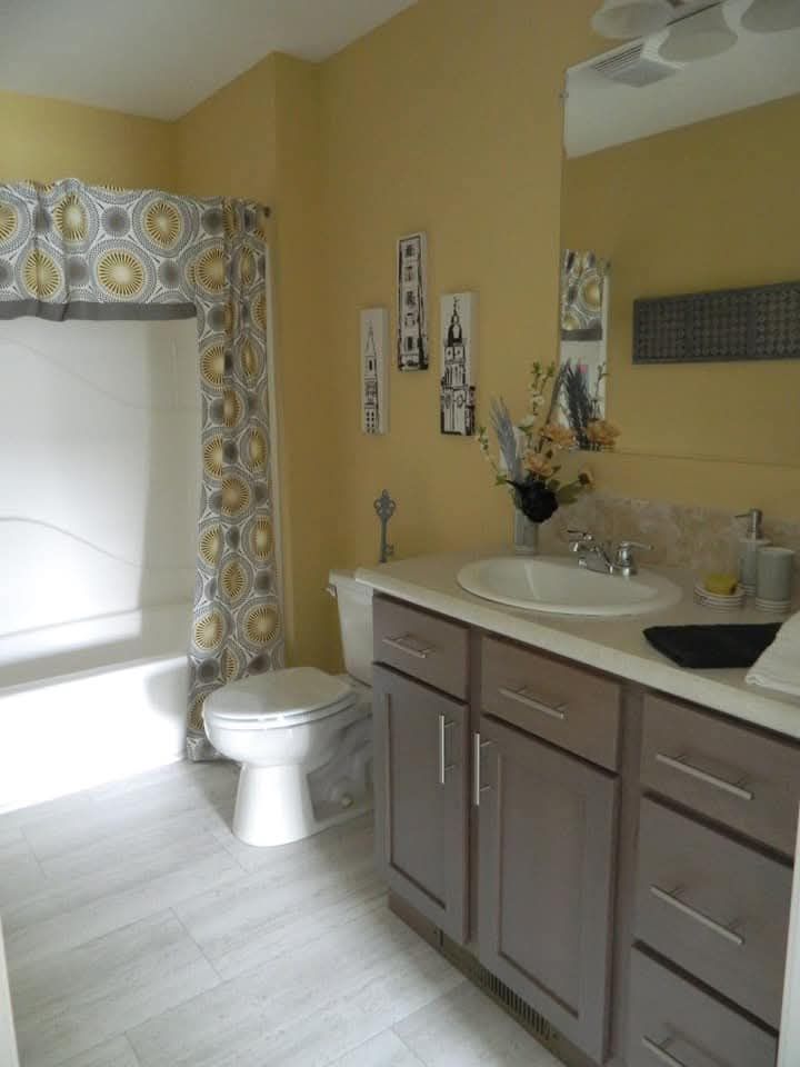 A small bathroom with light yellow walls features a gray vanity and sink. A patterned shower curtain with yellow accents hangs by the bathtub. Decor includes floral arrangements and wall art, creating a cozy atmosphere.