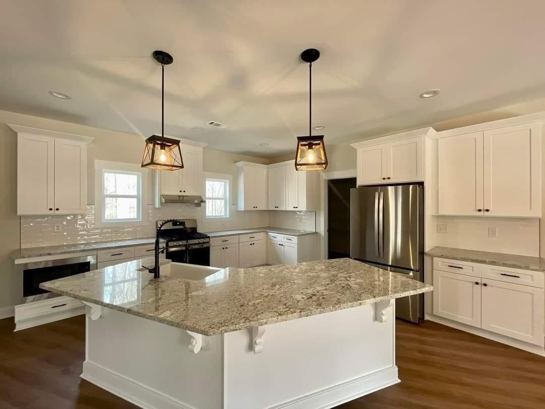Spacious kitchen with white cabinets, a large granite island, stainless steel fridge, dark wood floor, pendant lights, and bright natural light.