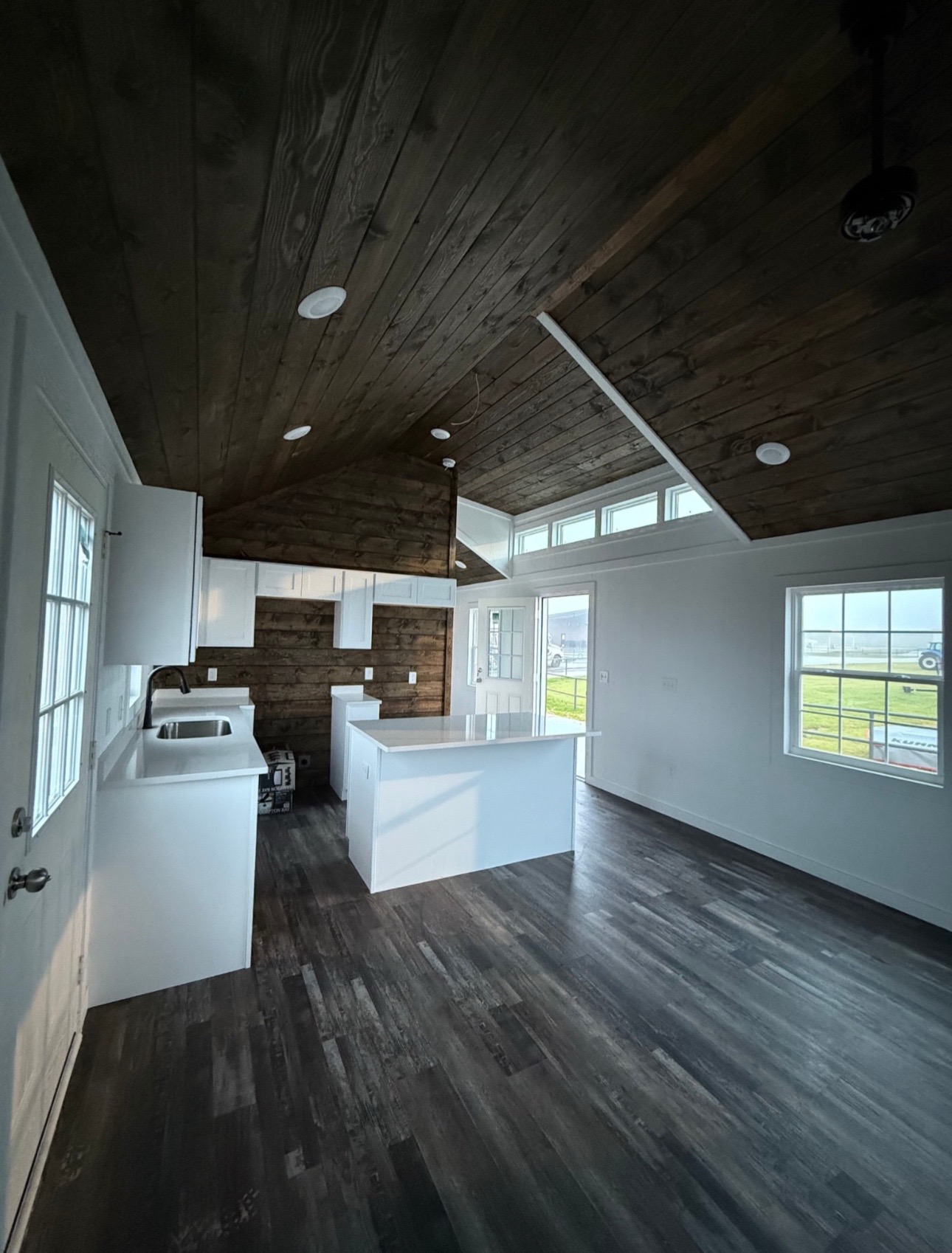 Modern, bright kitchen with dark wood ceiling and floor, white cabinets, and countertops. Large windows and glass door invite natural light. Cozy and inviting space.