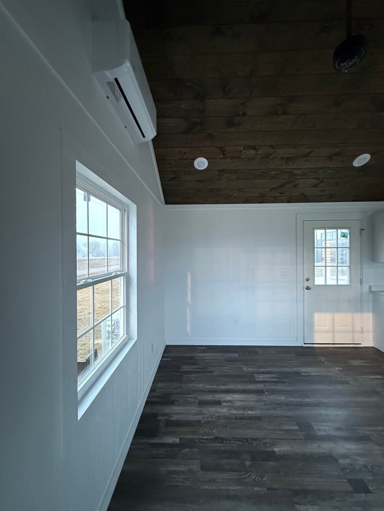 A small rustic room with a wooden ceiling and dark wooden floor, lit by natural light through a window and glass door. Calm, minimalist atmosphere.