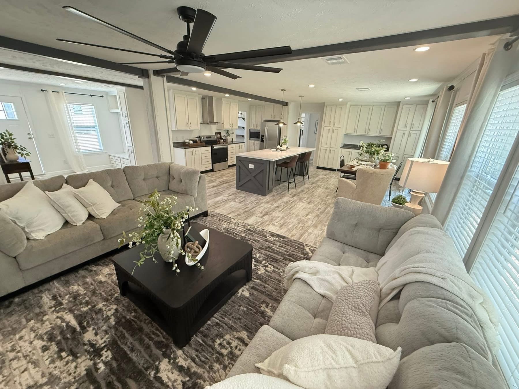 Spacious, modern living room and kitchen with gray sofas, white pillows, and cozy decor. Open kitchen features an island, pendant lights, and soft lighting.