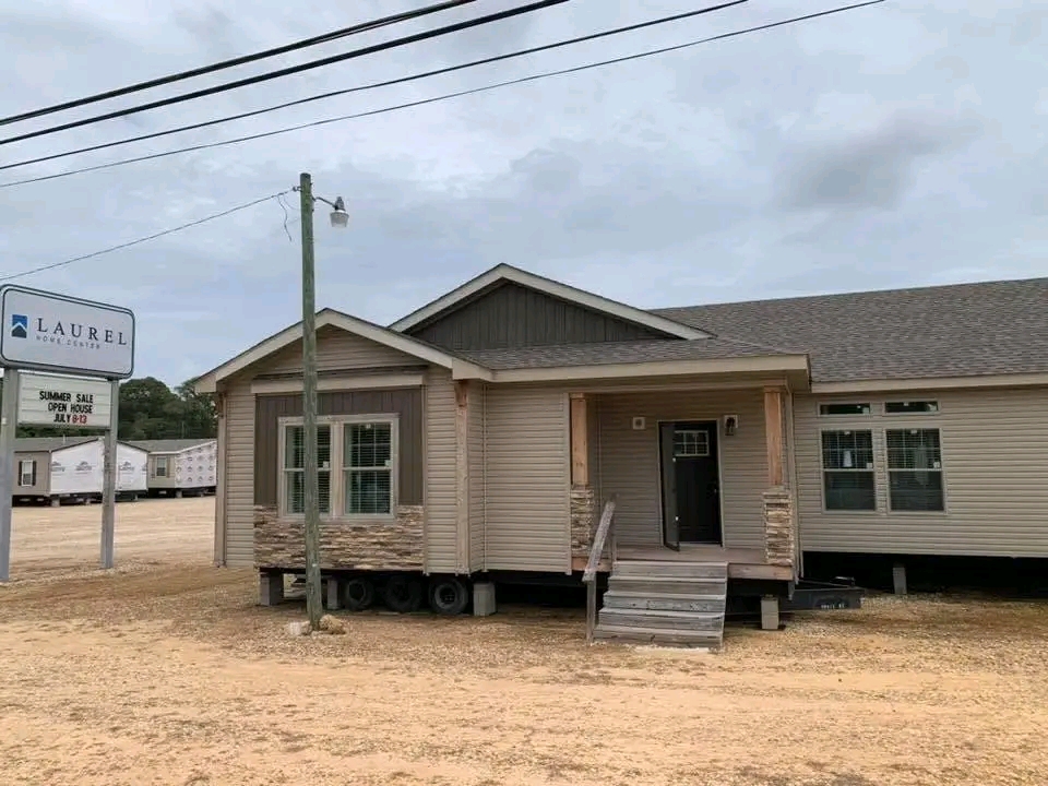 A beige modular home with stone accents is shown on a dirt lot. A "Laurel Home Center" sign is visible. The scene is overcast and calm.