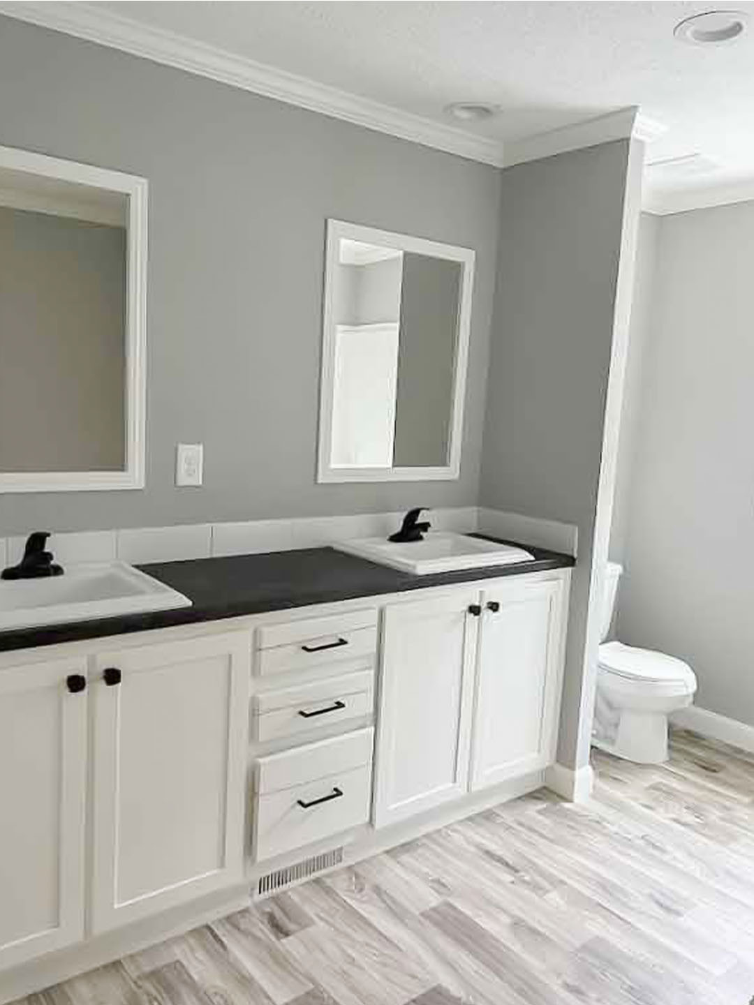 Modern bathroom with gray walls, a white double vanity, black faucets, and mirrors. Wood-look flooring, soft lighting, and a visible toilet convey a pristine, minimalist style.