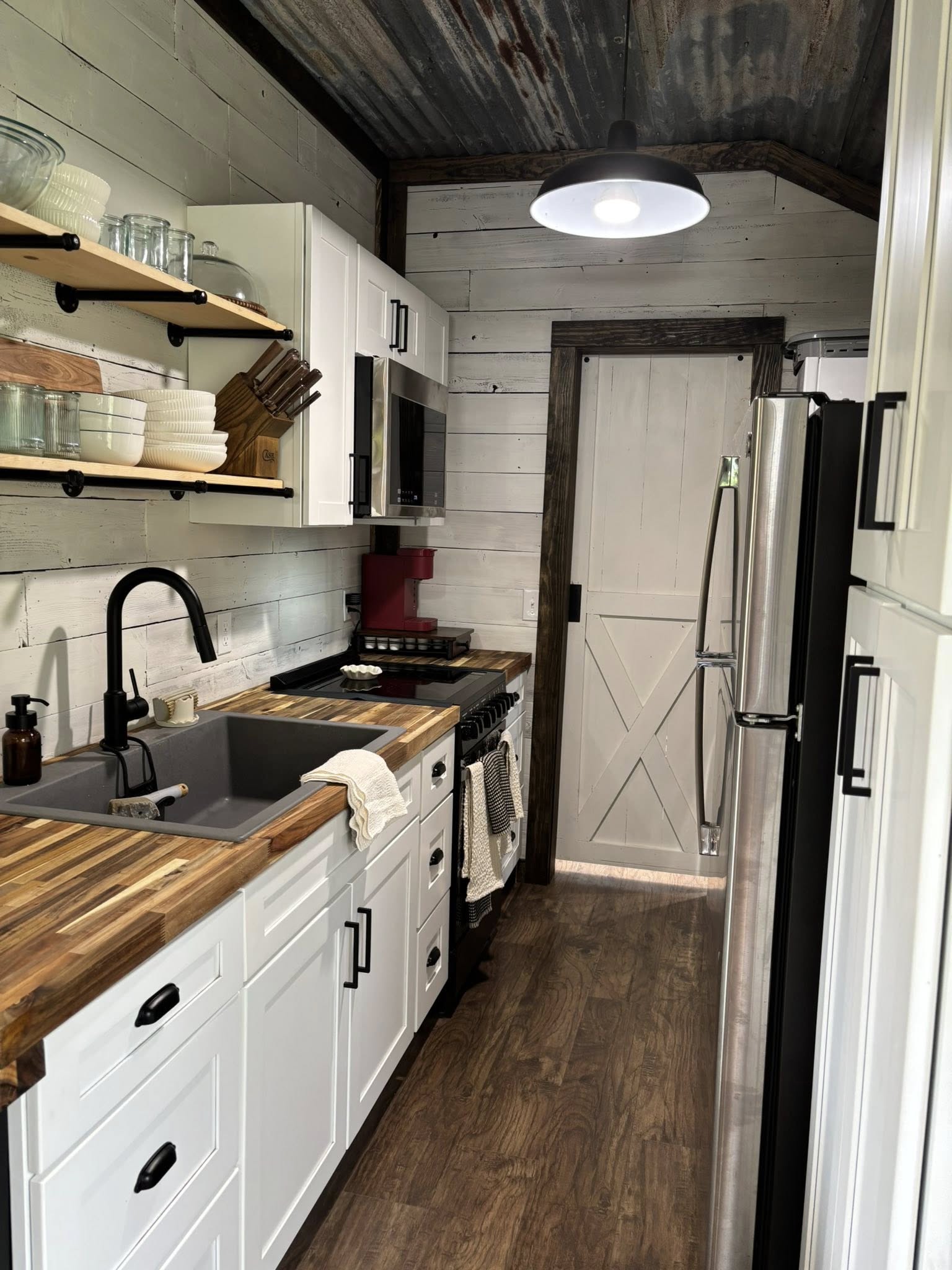 Rustic kitchen with white shiplap walls and a tin ceiling. Features include wooden countertops, open shelving, a black faucet, and stainless steel appliances.