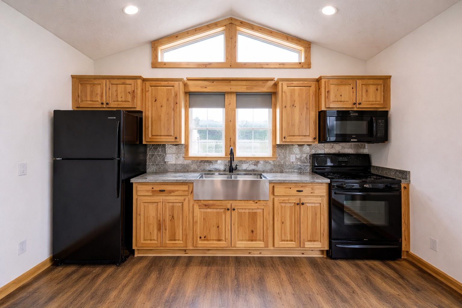 Rustic kitchen with wooden cabinets, stainless steel sink, black appliances, and a large central window. Warm, inviting tone with wooden flooring.