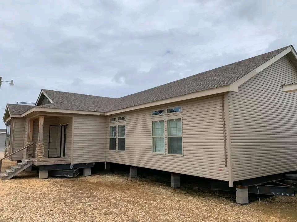 A beige modular home on cinder blocks, with a small wooden porch and steps. Overcast sky above, creating a neutral and calm atmosphere.