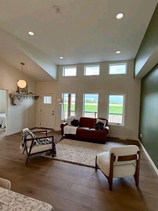 Spacious, modern living room with large windows overlooking greenery. Features a red sofa, white armchairs, wooden floor, and soft lighting. Calm ambiance.