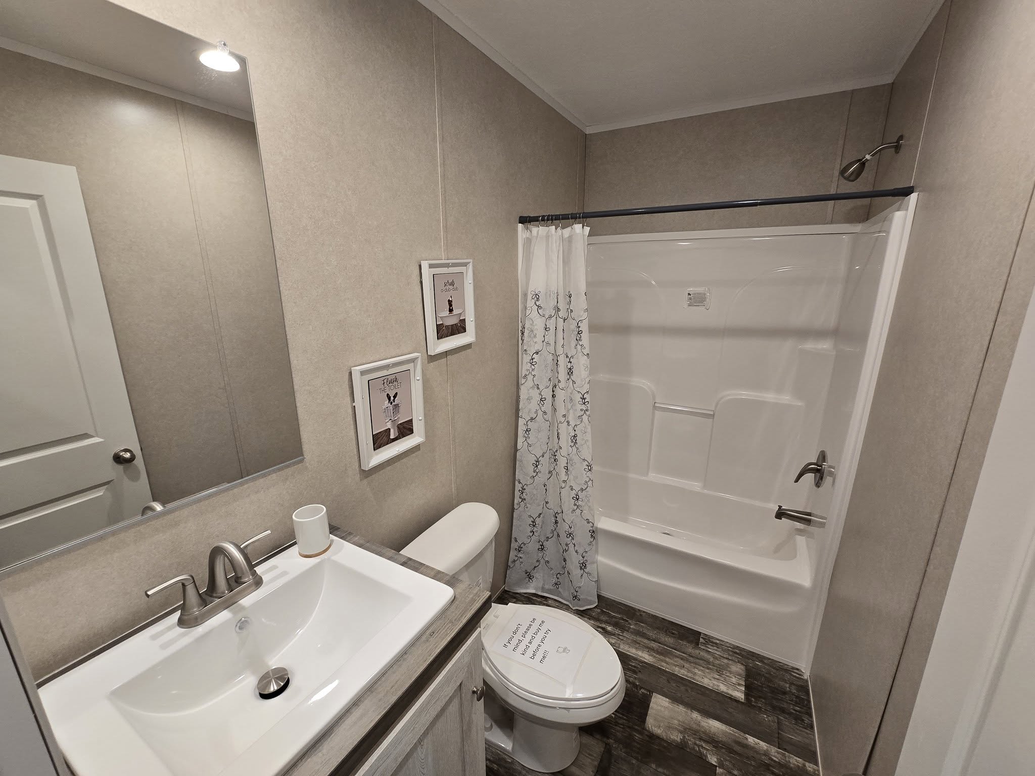 Small, modern bathroom with a shower-tub combo, white sink, and toilet. Beige walls, floral shower curtain, two framed pictures, and wood-look floor.