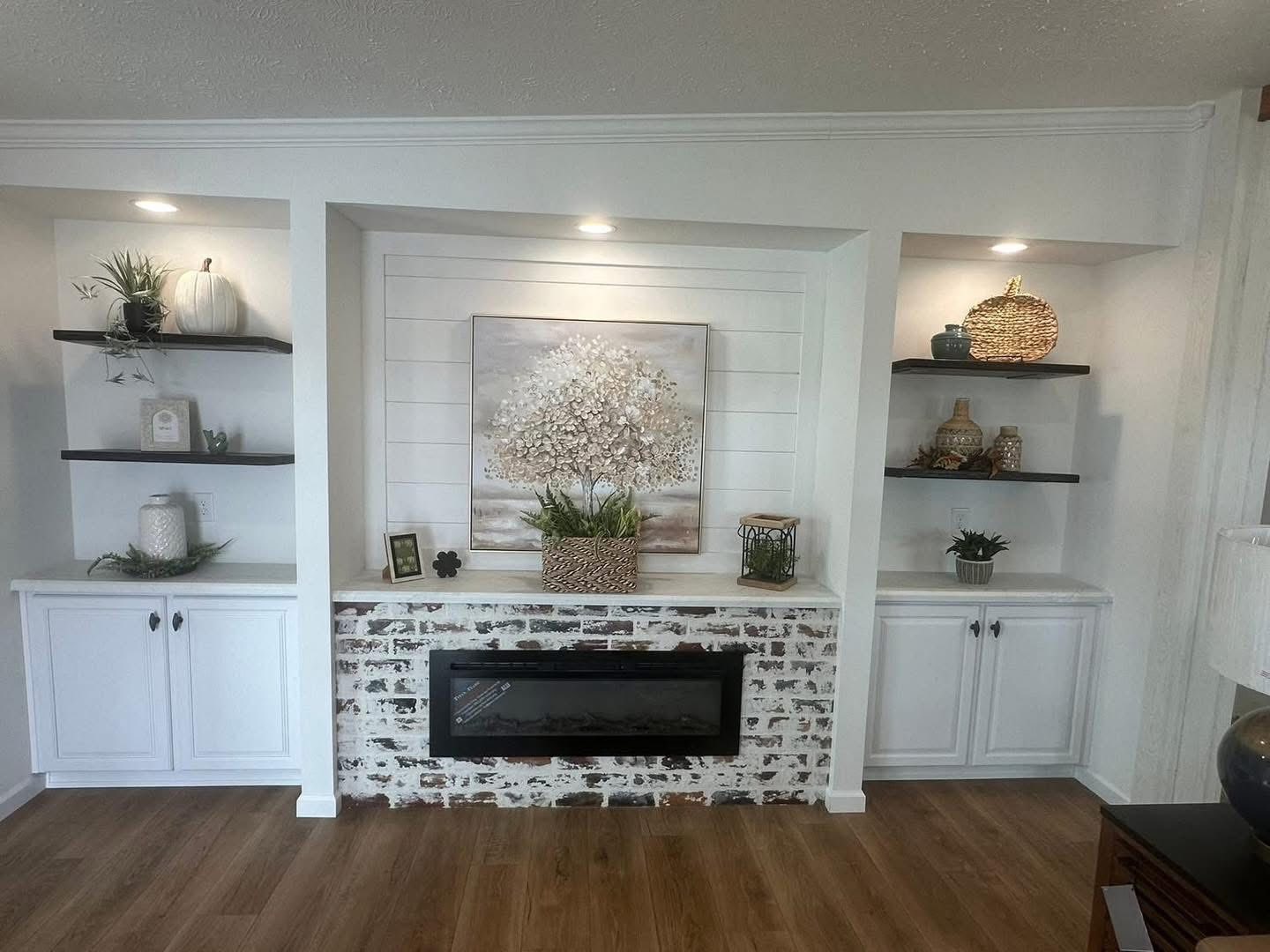 Cozy living room with a rustic brick fireplace and an elegant tree painting above. White shelves display decorative items, creating a serene ambiance.