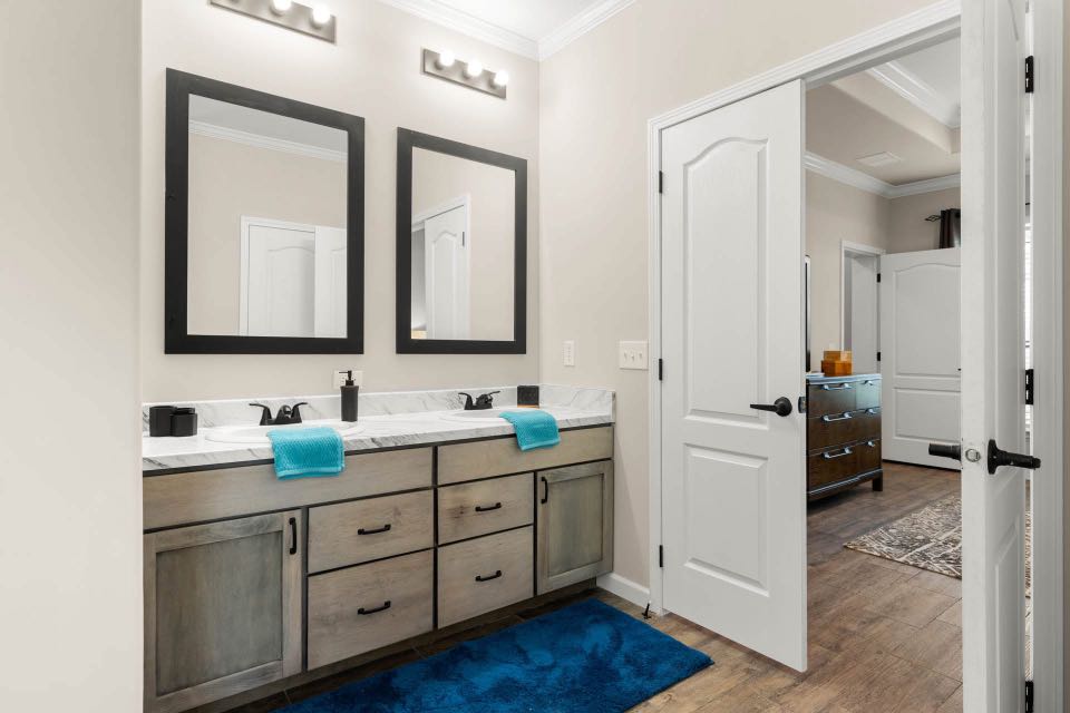 Spacious bathroom with double vanity, marble countertop, and two mirrors. Features modern light fixtures, blue accents, and open door revealing a bedroom.