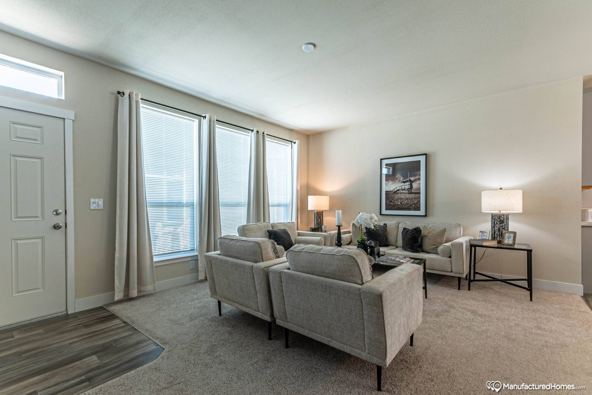 Bright living room with beige walls and carpet, featuring two sofas, two armchairs, side tables with lamps, and framed art. Large windows with curtains.
