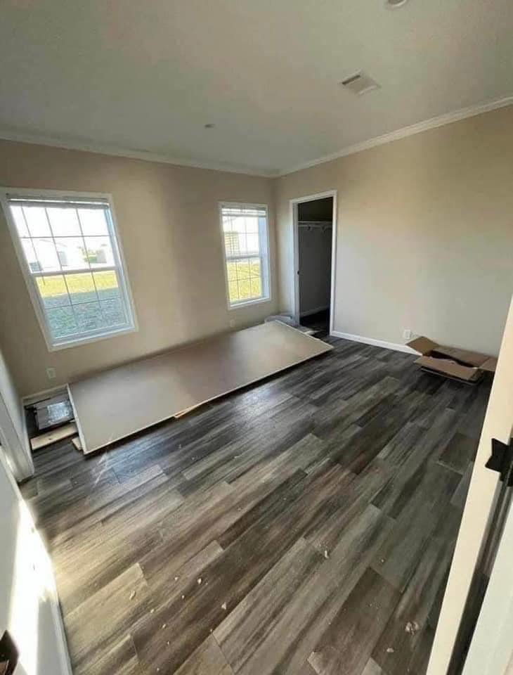 A spacious, light-filled room with two large windows and gray wood flooring. A detached door panel lies inside, leaning against a beige wall.