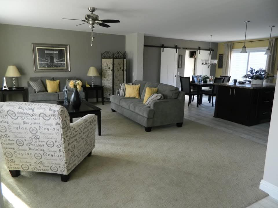 Spacious living room with gray sofas, yellow cushions, and a patterned armchair on a beige carpet. Open dining area and kitchen with soft lighting. Cozy, inviting tone.