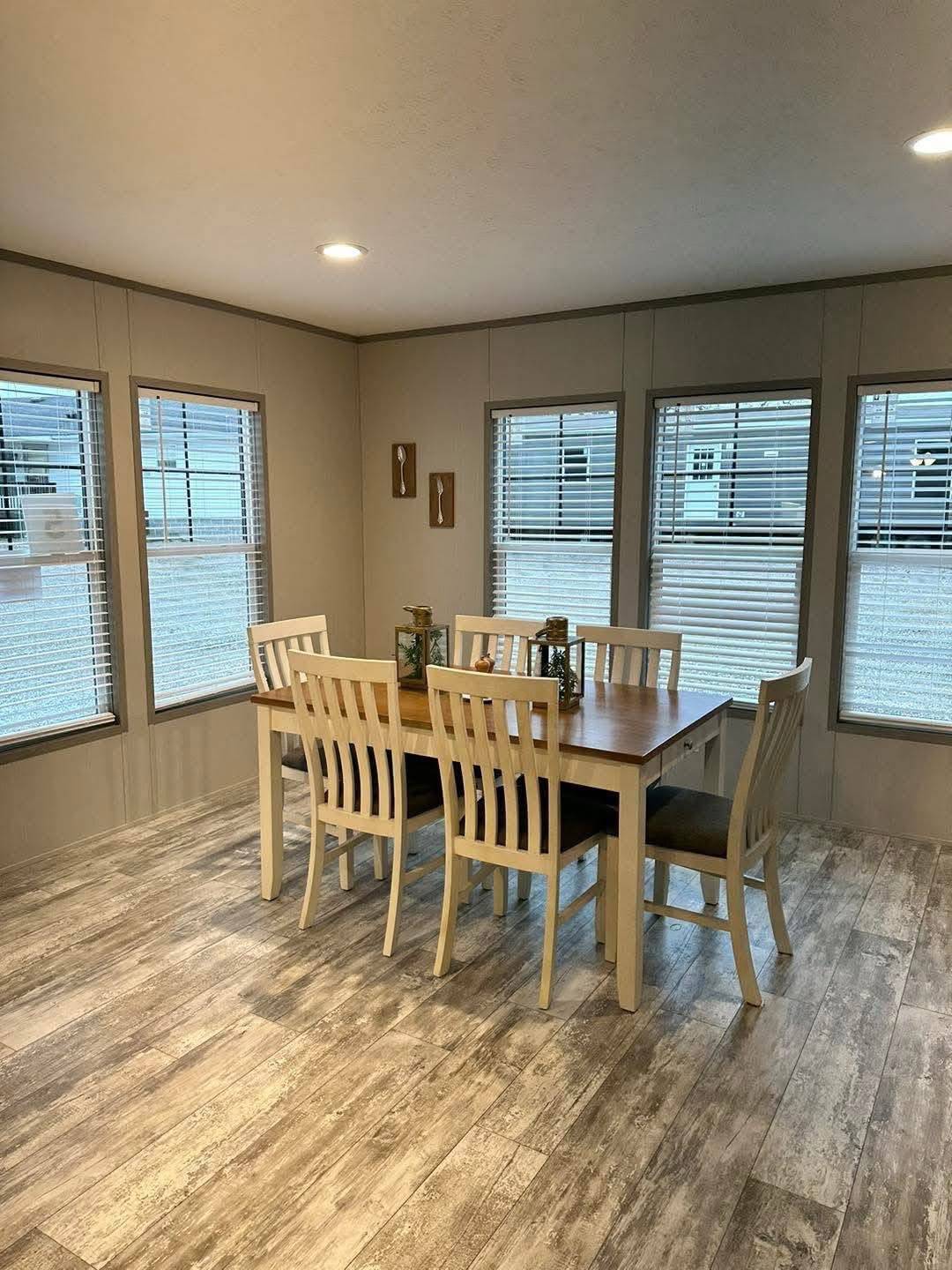 Bright dining room with a wooden table set for six, surrounded by large windows with blinds. The floor has a rustic wooden finish, creating a cozy ambiance.