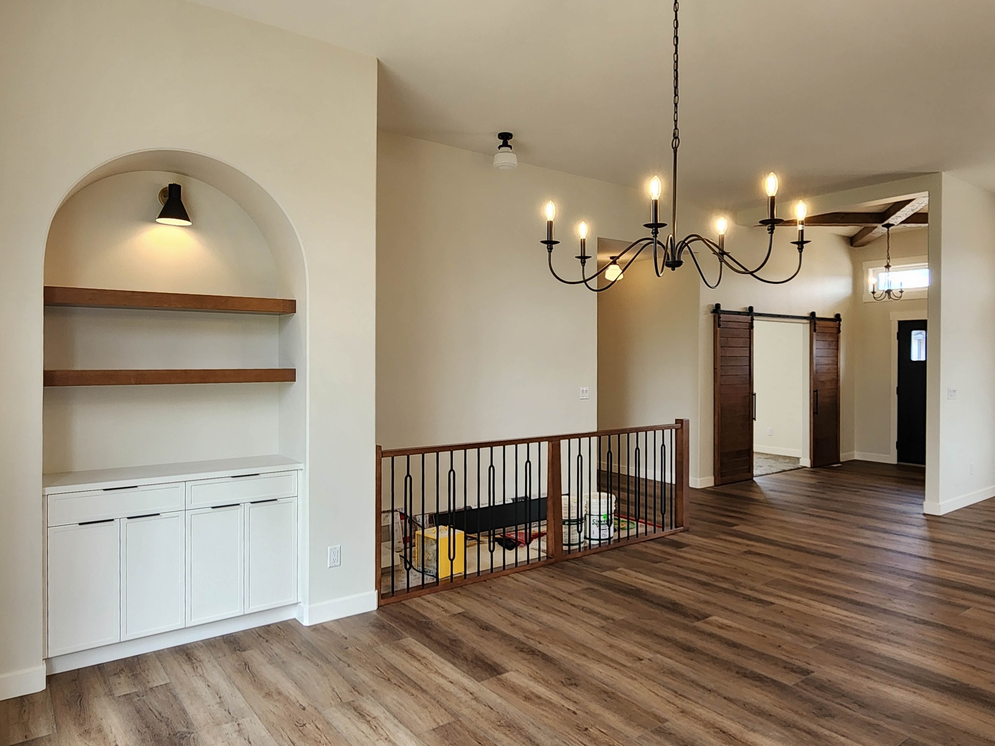 Spacious living area with light wood flooring, an empty arched bookshelf with a light, a large chandelier, and sliding barn doors in a cozy ambiance.