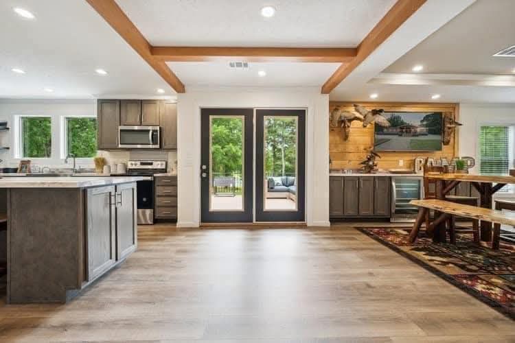 Spacious open kitchen-dining area with wooden beams, large French doors, modern gray cabinetry, and a dining table on a patterned rug.